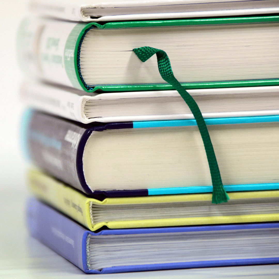 Image shows a pile of hardback books, they are blue, yellow, white and green with white pages. One has a dark green ribbon bookmark visible.