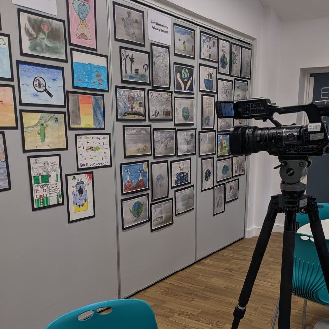 Image shows a wall covered in A4 pages of artwork with black boarders. In front is a television camera recording the images