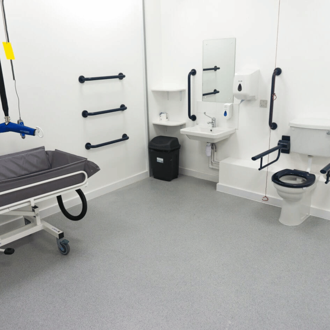 image shows a white room with a grey floor with a toilet, wash hand basin, grab rails and adult sized changing bench with a hoist above it.